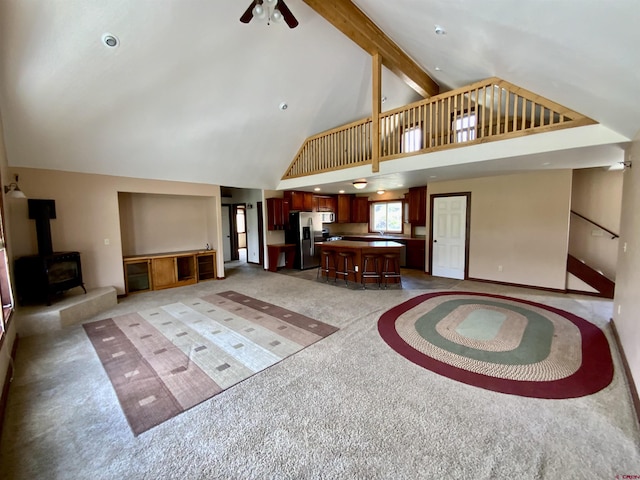 unfurnished living room featuring a wood stove, light carpet, high vaulted ceiling, ceiling fan, and beam ceiling