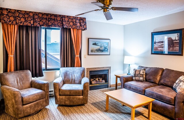 living area featuring a baseboard heating unit, a textured ceiling, a glass covered fireplace, and a ceiling fan