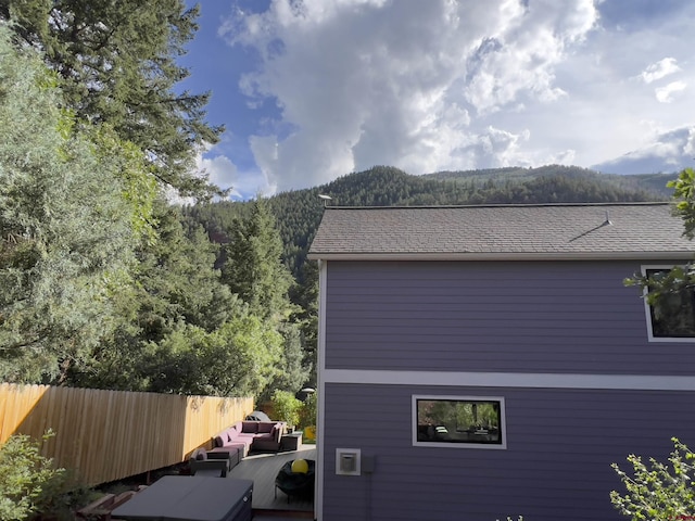 view of side of home with a shingled roof, a forest view, fence, an outdoor hangout area, and a mountain view