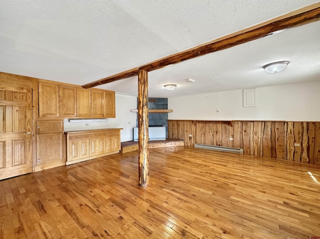 unfurnished living room with a textured ceiling, a baseboard heating unit, light wood-type flooring, wainscoting, and beamed ceiling