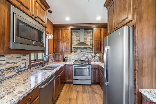 kitchen featuring wall chimney exhaust hood, appliances with stainless steel finishes, hanging light fixtures, light stone counters, and light wood-type flooring