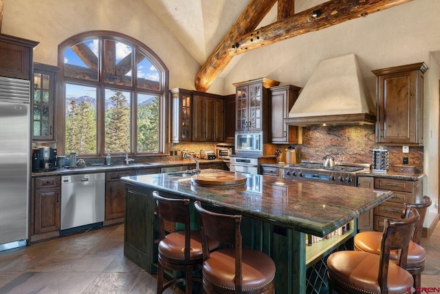 kitchen featuring an island with sink, beamed ceiling, a breakfast bar area, custom exhaust hood, and built in appliances