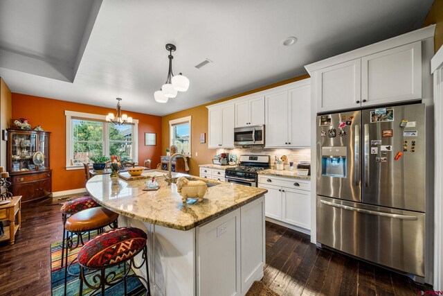 kitchen with appliances with stainless steel finishes, white cabinetry, an island with sink, light stone counters, and decorative light fixtures