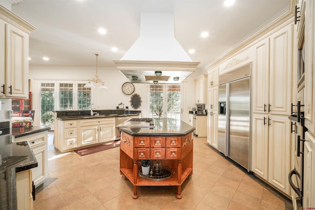 kitchen featuring an island with sink, premium range hood, cream cabinets, built in fridge, and decorative light fixtures