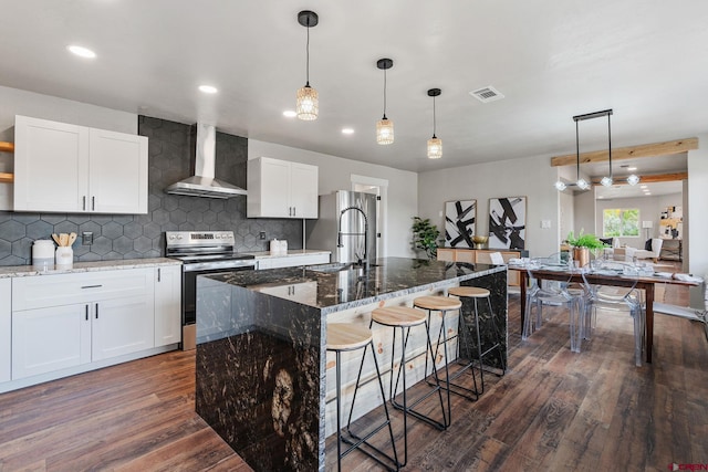 kitchen with wall chimney range hood, hanging light fixtures, a center island with sink, appliances with stainless steel finishes, and dark hardwood / wood-style flooring