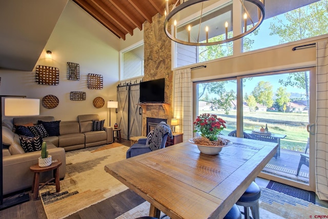 dining room featuring a stone fireplace, wood-type flooring, a barn door, a notable chandelier, and high vaulted ceiling