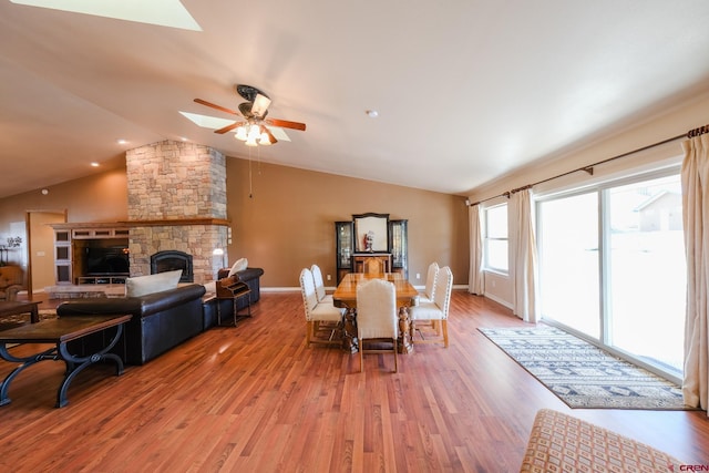 dining room with lofted ceiling with skylight, light hardwood / wood-style flooring, a stone fireplace, and ceiling fan