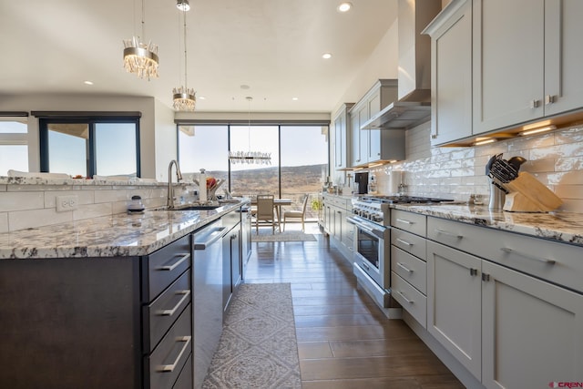 kitchen with stainless steel appliances, light stone countertops, decorative light fixtures, a kitchen island with sink, and wall chimney range hood