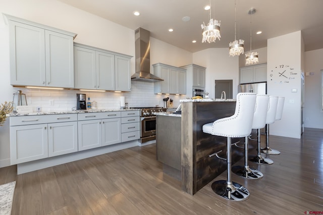 kitchen with stainless steel appliances, dark wood-type flooring, light stone counters, wall chimney range hood, and decorative light fixtures