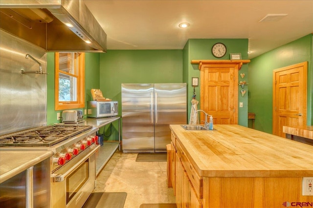 kitchen with sink, an island with sink, range hood, appliances with stainless steel finishes, and butcher block countertops