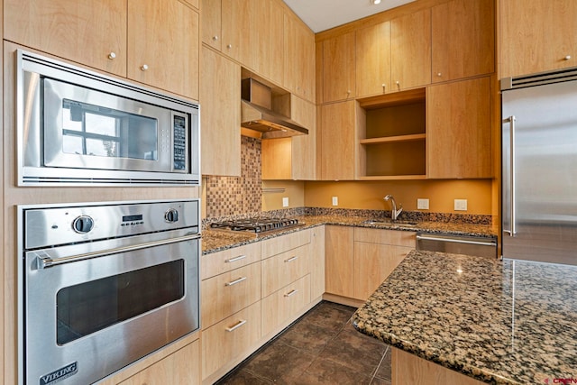 kitchen with light brown cabinets, dark stone counters, sink, built in appliances, and wall chimney exhaust hood