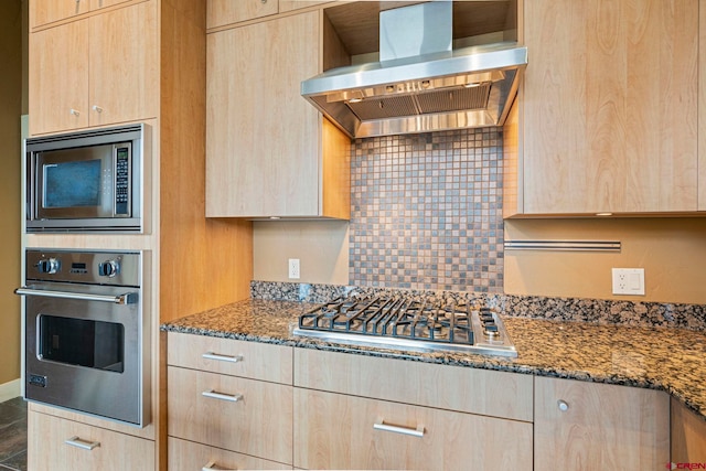 kitchen featuring backsplash, dark stone counters, stainless steel appliances, wall chimney range hood, and light brown cabinets