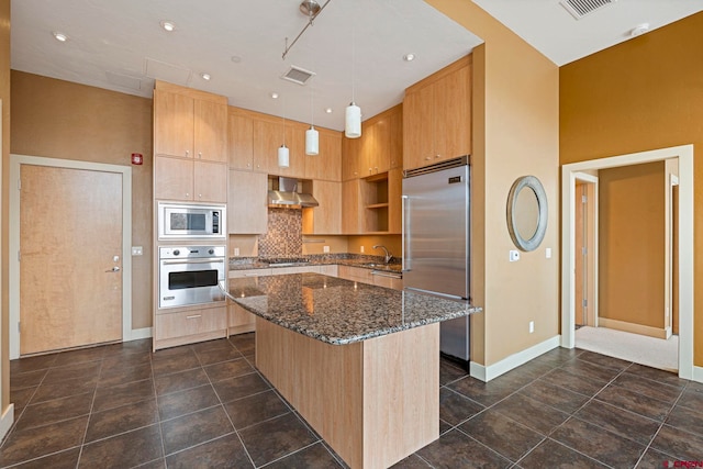 kitchen with dark stone counters, wall chimney exhaust hood, built in appliances, decorative light fixtures, and a center island