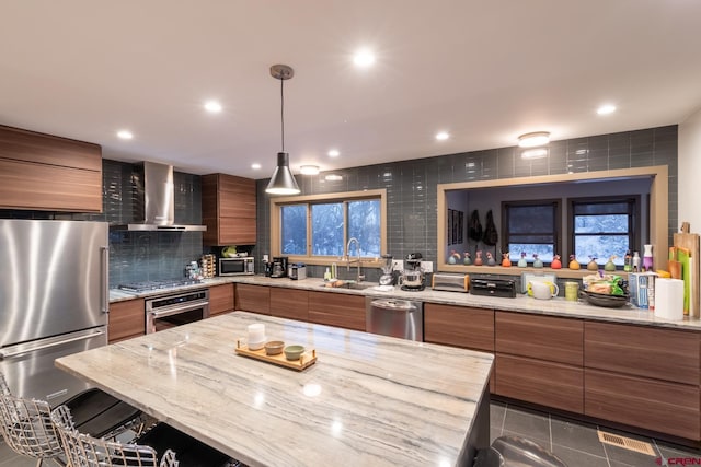 kitchen with light stone countertops, appliances with stainless steel finishes, wall chimney exhaust hood, sink, and hanging light fixtures