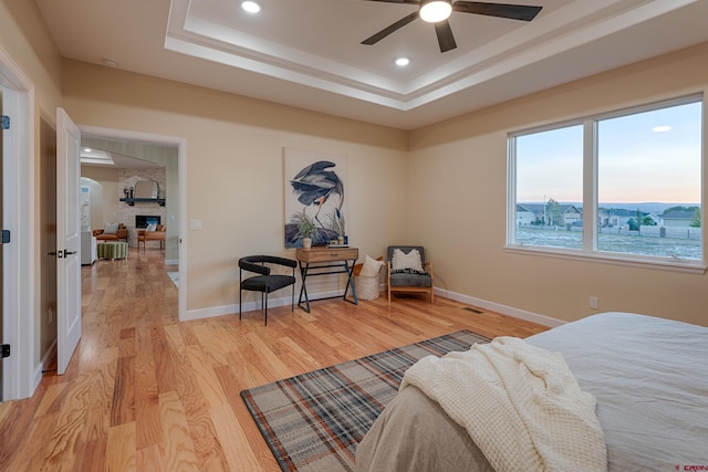 bedroom with light wood-type flooring, a raised ceiling, ceiling fan, and a stone fireplace