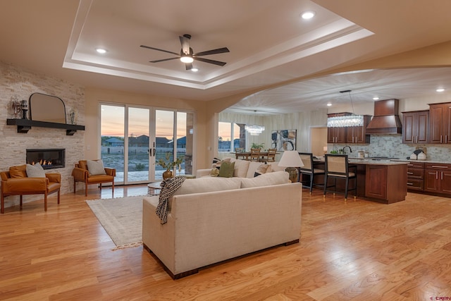 living room featuring a tray ceiling, ceiling fan, sink, light hardwood / wood-style flooring, and a fireplace