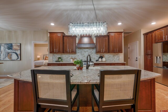 kitchen featuring stainless steel fridge, custom exhaust hood, a center island with sink, a chandelier, and light hardwood / wood-style floors