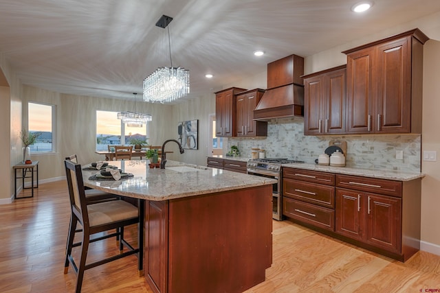 kitchen with sink, stainless steel gas range oven, premium range hood, decorative light fixtures, and a center island with sink