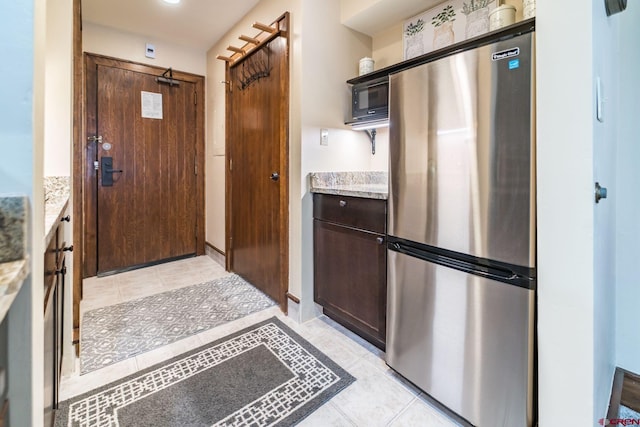 kitchen featuring light tile patterned floors, light stone counters, dark brown cabinetry, black microwave, and stainless steel refrigerator