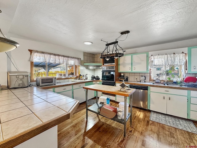 kitchen with tile countertops, backsplash, hanging light fixtures, stainless steel dishwasher, and light hardwood / wood-style floors