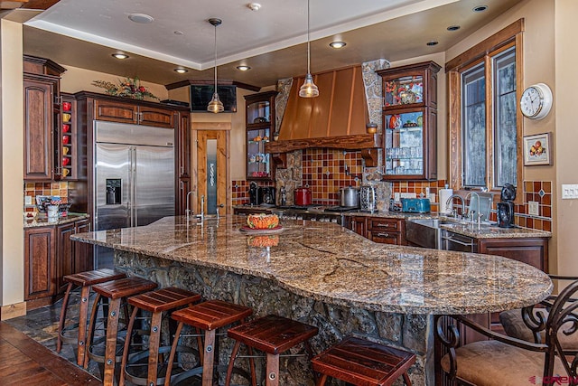 kitchen with pendant lighting, custom exhaust hood, sink, a kitchen island, and stainless steel built in refrigerator