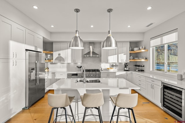 kitchen featuring a kitchen island with sink, wall chimney range hood, appliances with stainless steel finishes, white cabinetry, and beverage cooler