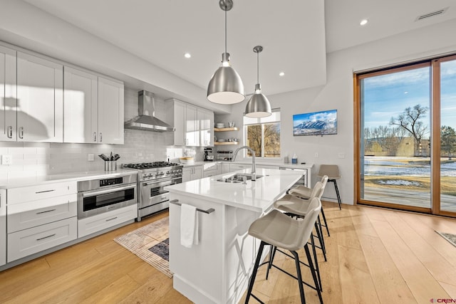 kitchen with appliances with stainless steel finishes, a kitchen island with sink, wall chimney range hood, decorative light fixtures, and white cabinets