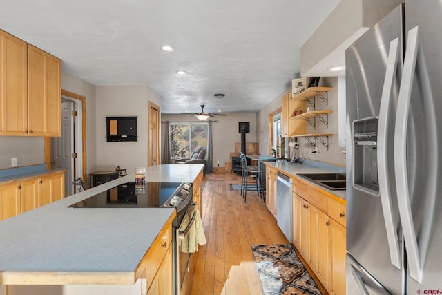 kitchen featuring a wood stove, ceiling fan, a center island, light brown cabinetry, and appliances with stainless steel finishes