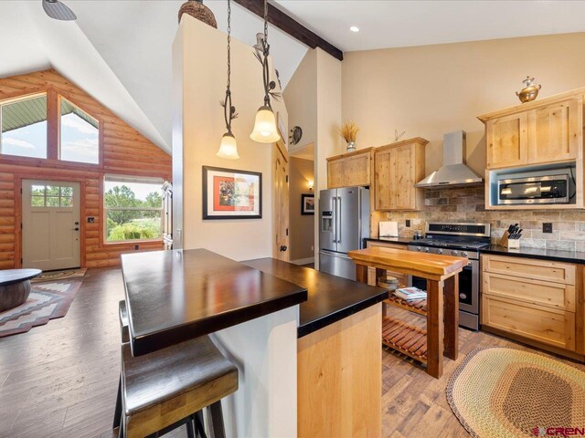 kitchen with light brown cabinetry, backsplash, wall chimney exhaust hood, stainless steel appliances, and hanging light fixtures