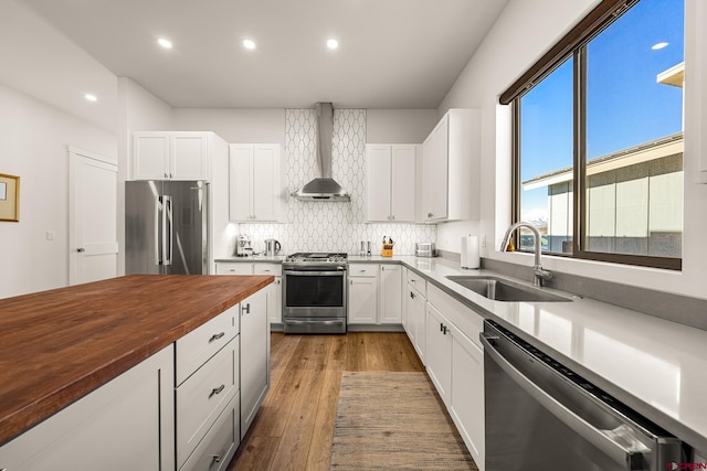 kitchen with appliances with stainless steel finishes, sink, wall chimney range hood, white cabinetry, and butcher block counters