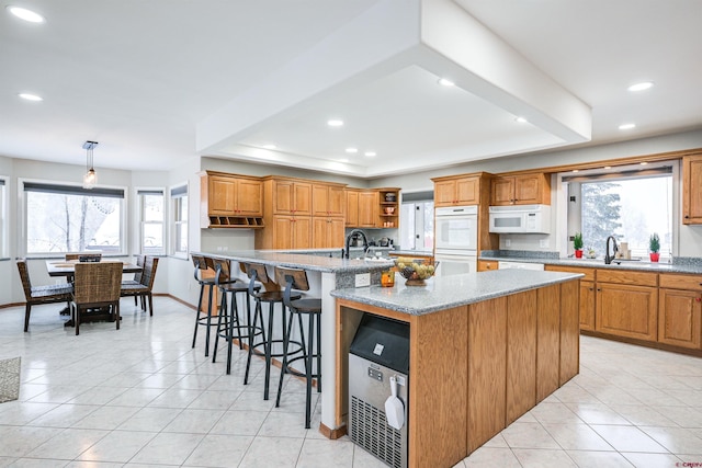 kitchen with hanging light fixtures, a raised ceiling, an island with sink, white appliances, and a breakfast bar area