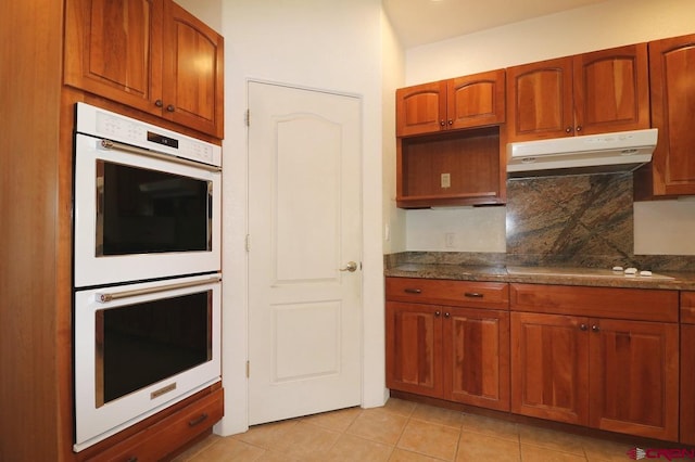 kitchen featuring backsplash, light tile patterned flooring, dark stone counters, and white appliances