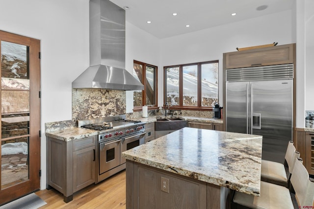 kitchen featuring sink, wall chimney exhaust hood, premium appliances, light stone counters, and backsplash