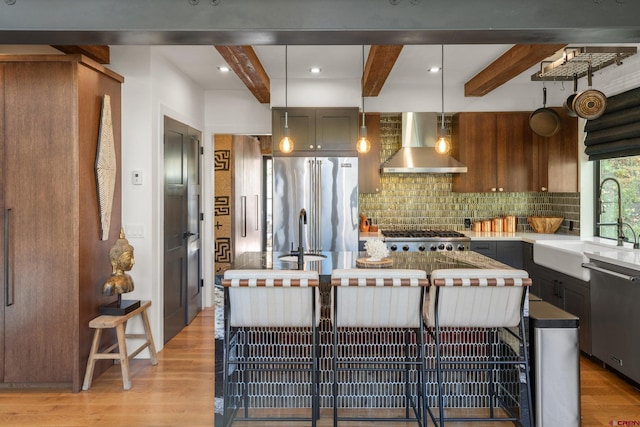 kitchen featuring appliances with stainless steel finishes, a center island, beamed ceiling, wall chimney range hood, and decorative light fixtures