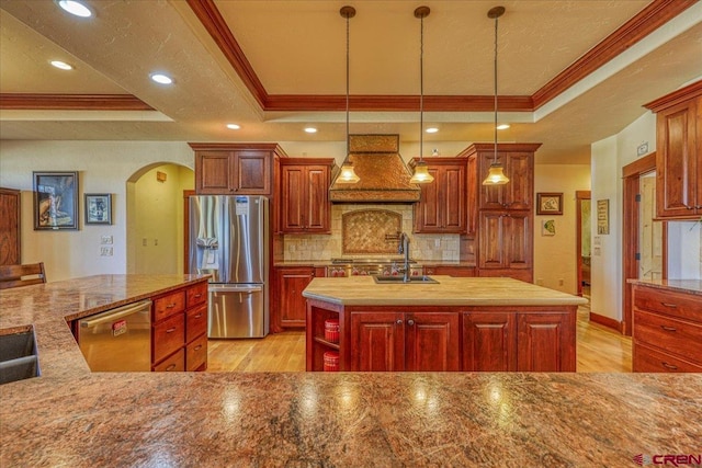 kitchen featuring a tray ceiling, appliances with stainless steel finishes, a center island with sink, and custom exhaust hood