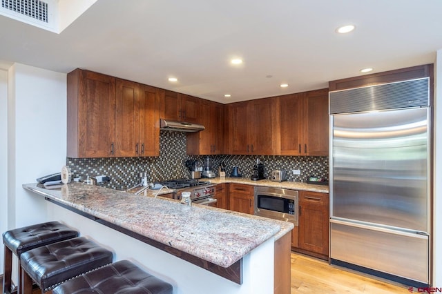 kitchen featuring light stone countertops, a kitchen bar, built in appliances, kitchen peninsula, and light hardwood / wood-style flooring