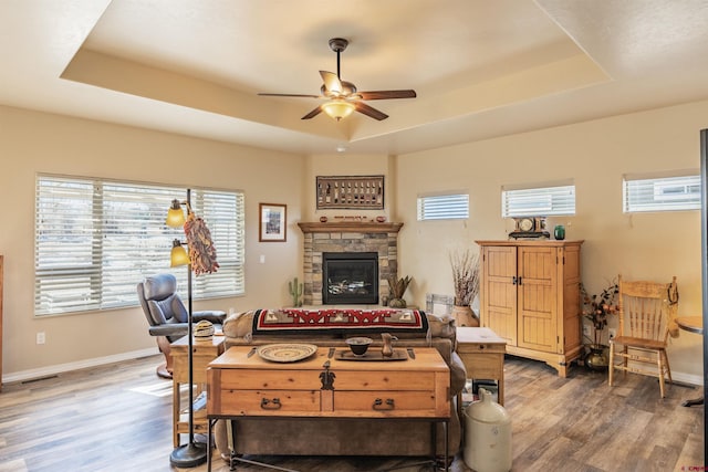 living room featuring dark wood-type flooring, ceiling fan, a tray ceiling, and a stone fireplace