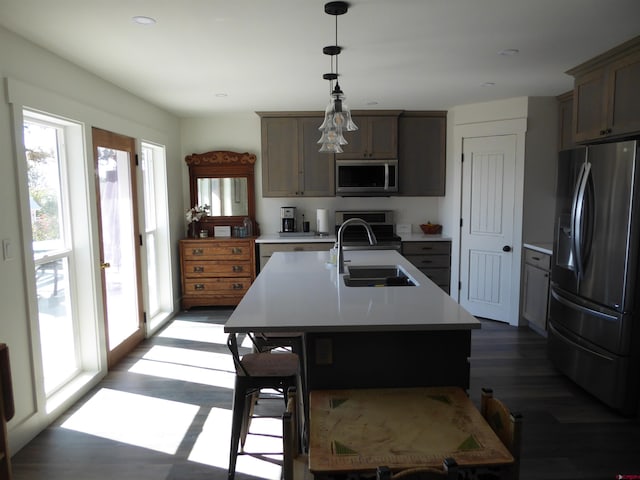 kitchen with an island with sink, sink, hanging light fixtures, stainless steel appliances, and dark wood-type flooring