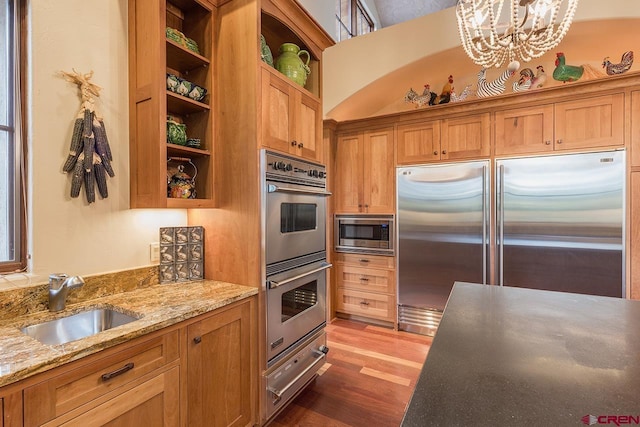 kitchen with sink, dark wood-type flooring, built in appliances, light stone countertops, and decorative light fixtures