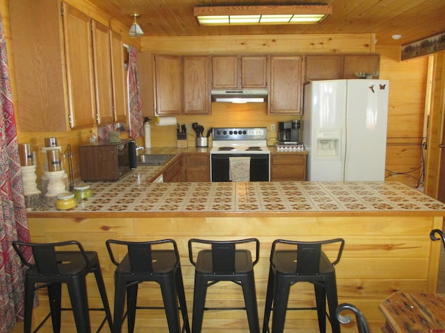 kitchen featuring tile countertops, wood walls, a kitchen breakfast bar, wood ceiling, and white appliances