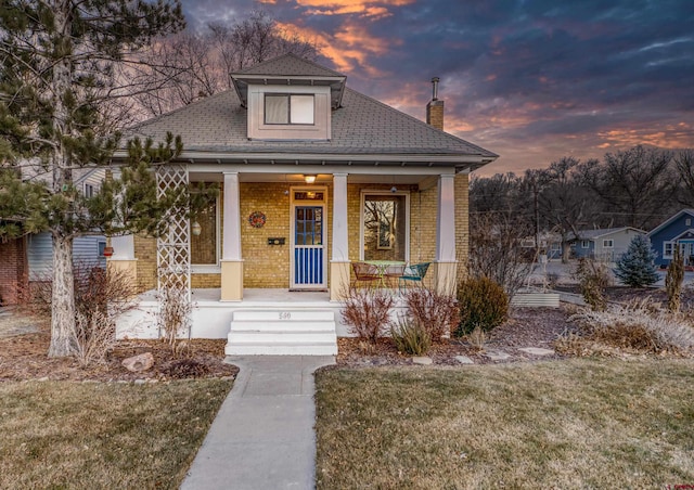 bungalow with covered porch, brick siding, a chimney, and a front lawn
