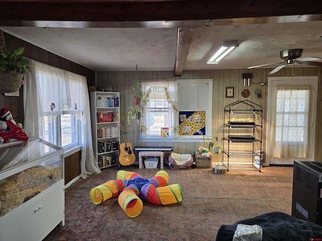 recreation room with beamed ceiling, wooden walls, a healthy amount of sunlight, and carpet flooring