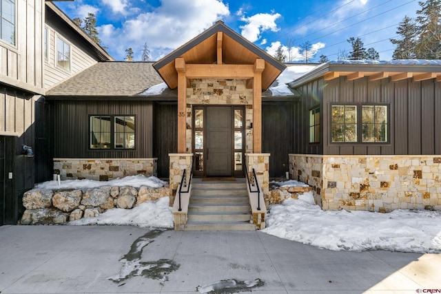 snow covered property entrance featuring board and batten siding, stone siding, and a shingled roof