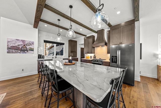 kitchen featuring light stone counters, a kitchen island, stainless steel refrigerator with ice dispenser, custom exhaust hood, and range with gas cooktop