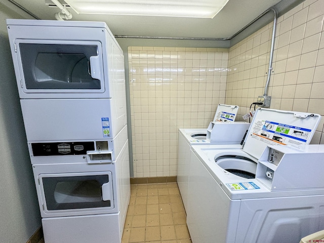 community laundry room with stacked washing maching and dryer, washing machine and dryer, light tile patterned floors, and tile walls