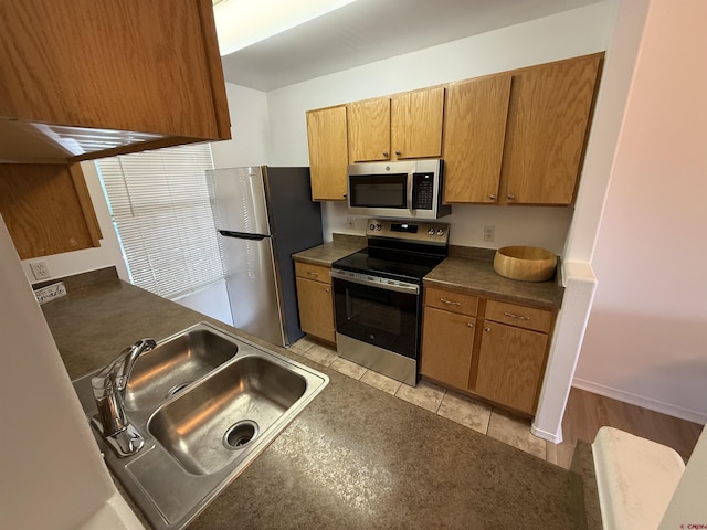 kitchen with dark countertops, appliances with stainless steel finishes, brown cabinetry, light tile patterned flooring, and a sink