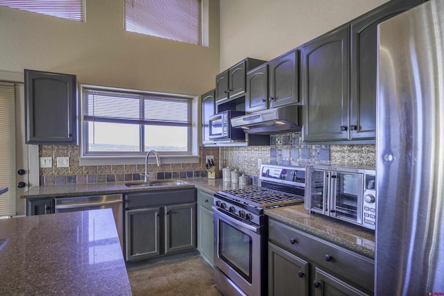 kitchen with dark stone counters, a sink, stainless steel appliances, under cabinet range hood, and backsplash