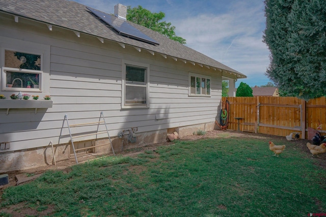 view of side of home with a chimney, a shingled roof, a lawn, roof mounted solar panels, and fence