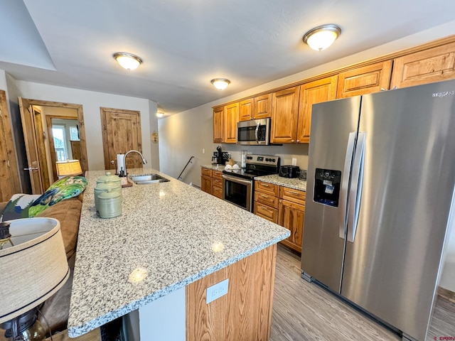 kitchen with light wood finished floors, brown cabinetry, appliances with stainless steel finishes, light stone countertops, and a sink