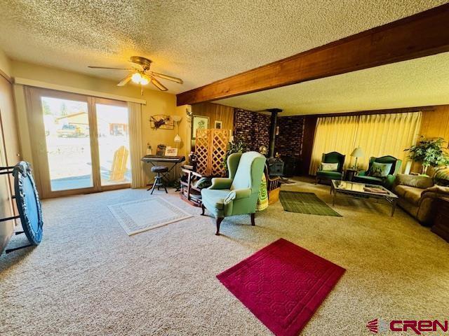 living area with carpet floors, beam ceiling, a ceiling fan, a wood stove, and a textured ceiling